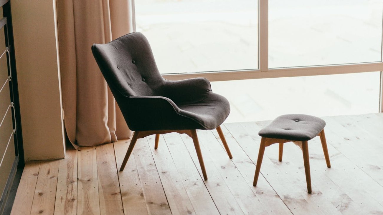 A contemporary armchair and footstool in a minimalistic room with sunlight streaming through large windows.