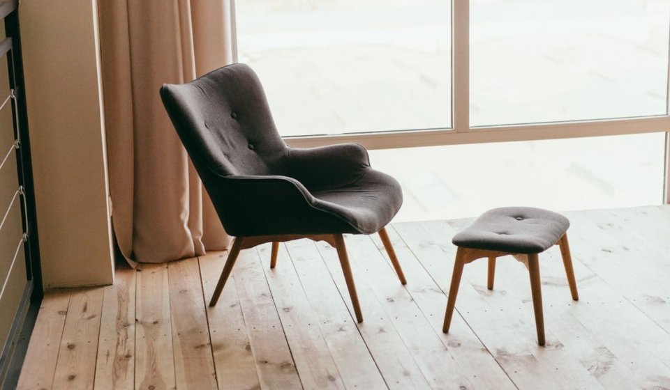 A contemporary armchair and footstool in a minimalistic room with sunlight streaming through large windows.